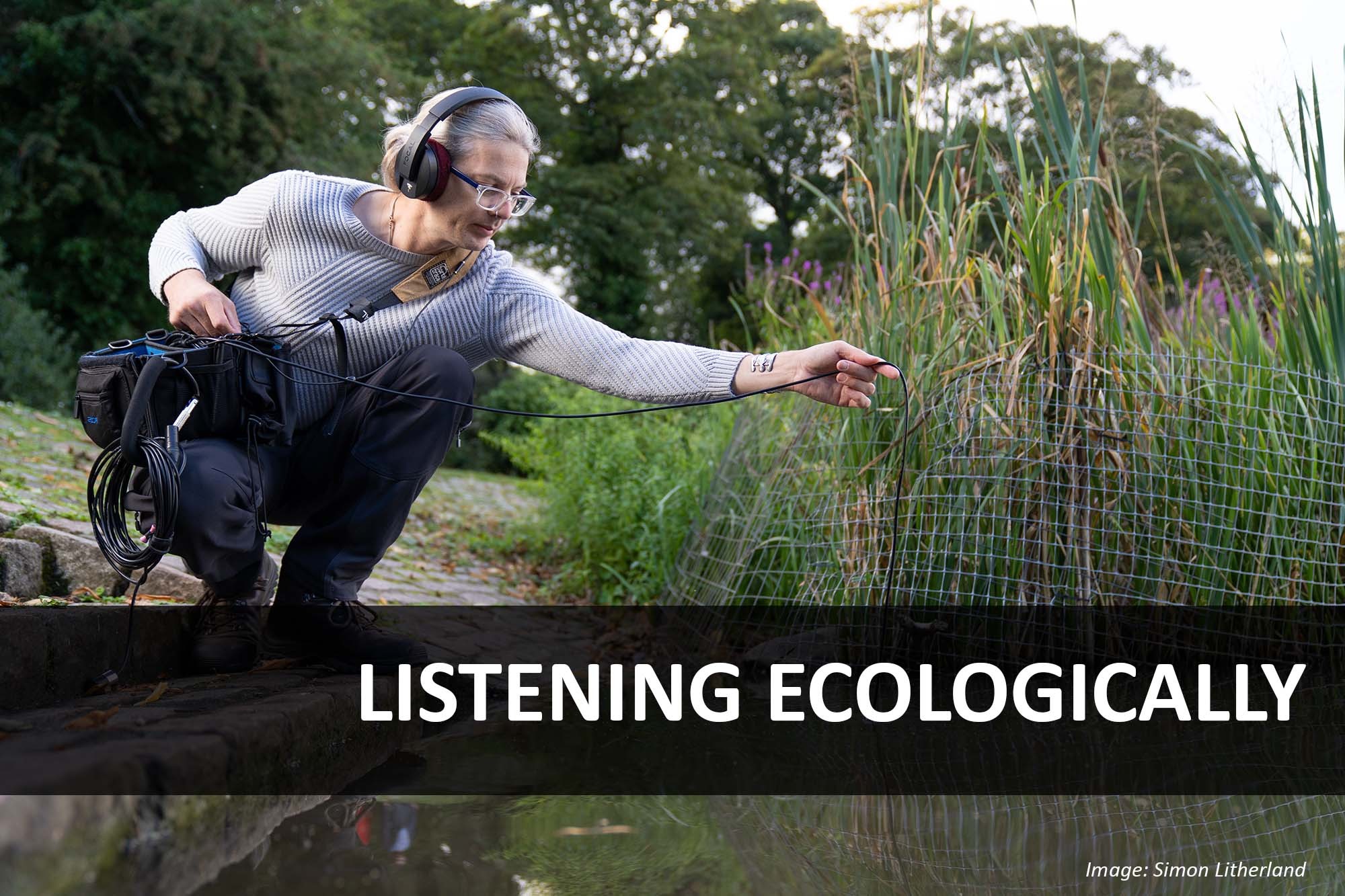 A man wearing headphones crouches on steps leading down to water and lowers a microphone below the surface next to the water reeds. Text reads 'Listening Ecologically'